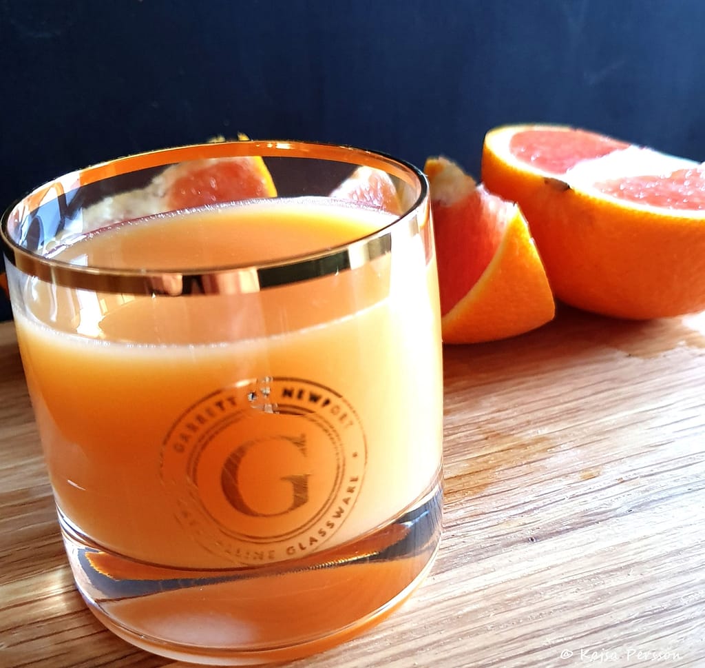 Litet glas med guldkant och guldekor med orangea juice på en skärbräda. Bakom ligger en delad blodapelsin i klyftor