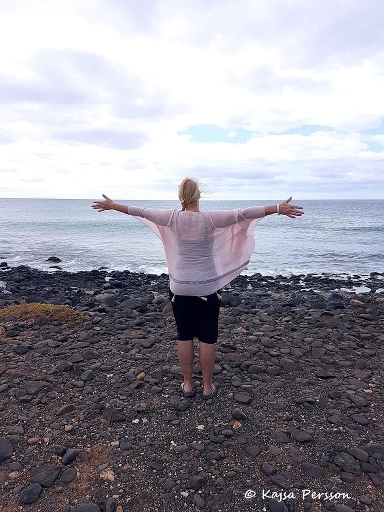 Kvinna vänd mot havet, sträcker ut händerna åt sidan. Den rosa sjalen fladdrar i vinden.